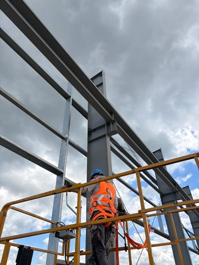 The image shows a construction worker wearing a blue hard hat and an orange safety vest, harnessed and standing on a yellow lift platform. The worker is on a construction site in Stróża, Poland, amidst a metal framework under a cloudy sky. The worker appears to be performing a task related to the construction of a large metal structure, possibly a building or a warehouse. The scene depicts a typical day on a construction site, with elements of heavy industry and personal safety equipment.