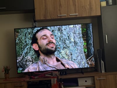 A television displays a scene with a bearded man in a plaid shirt leaning against a tree. The television sits on a wooden cabinet. Pink hair straighteners, hair dye and a desk calendar showing April 14 are on the cabinet. A Playstation 5 is next to the TV. The location is in a home; there's no specific evidence to show it is Kliment, Bulgaria.