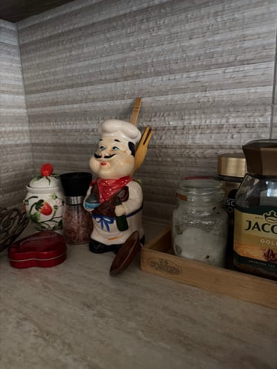 The image depicts a shelf in what appears to be a domestic kitchen setting, likely located in the city of Dromolaxia - Meneou Municipality, Cyprus. The shelf itself has a textured, horizontally-striped light gray background and a speckled beige countertop.
On the shelf, several objects are arranged. To the left, there is a white ceramic jar adorned with painted strawberries and a red lid topped with a red strawberry. Next to it is a clear glass salt or pepper grinder containing pink crystals.
Dominating the center of the shelf is a ceramic figurine of a chef, rendered in a cartoonish style. The chef wears a white chef's hat, a red bandana, and a white outfit with blue trim. He has a large mustache and is holding a bottle in his left hand and a utensil in his right, which is partially obscured.
To the right of the chef figurine, a clear glass jar filled with white granules and some darker specks is situated within a light-colored wooden tray. Behind this jar, a dark brown jar with a gold lid is visible, and further to the right, a glass jar of "Jacobs Gold" coffee is prominently displayed, with text indicating "SOURCING FOR E".
In the foreground, a red heart-shaped tin is visible on the left side of the shelf. A small, dark brown, spoon-shaped object rests on the shelf in front of the chef figurine. The overall arrangement suggests a collection of kitchen-related items, possibly decorative or functional.