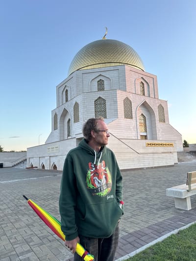 A man stands in front of a large white building with a golden dome, holding a closed, rainbow-colored umbrella. The man is wearing a green hoodie with a graphic print and glasses, and he is looking to his right. The building appears to be a mosque or a mausoleum, with arched windows and intricate stonework. The ground is paved with interlocking bricks, and there is a wooden bench visible to the right of the man. The sky is clear and blue, suggesting it is daytime. The location is the city of Bolgar, Russia, a UNESCO World Heritage site known for its historical Bulgarian architecture.