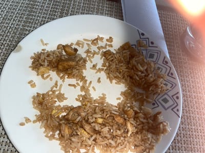 A partially eaten plate of fried rice rests on a woven placemat. A napkin sits to the left of the plate. The rice appears to contain chicken and egg. The plate is white with a geometric pattern on the rim.