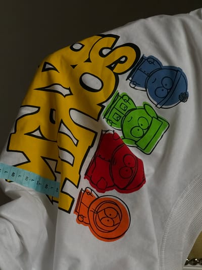 A white t-shirt featuring characters from the animated show "South Park" is laid out flat. The print includes four characters in bold outlines and vibrant colors: a blue character wearing a blue hat with a pom-pom, a green character in a green hat, a red character in a red hooded jacket, and an orange character in an orange hooded jacket. Below the characters, large yellow lettering with black outlines spells out "SOUTH." A blue price tag with "L" and "40" printed repeatedly is attached to the lower right side of the t-shirt. The background is dark and out of focus, suggesting the t-shirt is on display in a retail setting or being photographed. The lighting is artificial, and no specific time of day or weather conditions are indicated. The setting appears to be indoors, possibly a store. The characters' expressions are mostly neutral or slightly grumpy.