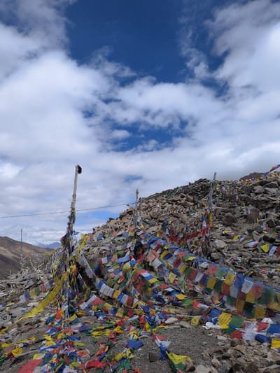 This image depicts a mountainous landscape near Sasoma, India, characterized by a vast expanse of colorful prayer flags. The flags are strung across poles and draped over rocks, creating a vibrant tapestry against the rugged terrain. In the background, snow-capped mountains are visible under a dramatic sky filled with white clouds against a deep blue. The ground is rocky and uneven, with loose stones and gravel. The overall scene suggests a high-altitude, spiritual location.