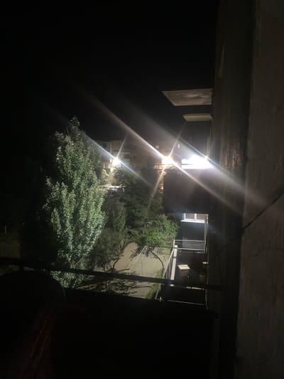 In Kemin, Kyrgyzstan, this nighttime scene captures an urban environment illuminated by streetlights. A tall, leafy tree dominates the left side of the frame, its foliage catching the bright light from a powerful lamp positioned on a building in the background. The building itself appears to be an apartment complex, with multiple windows visible. Below the tree, a road winds through the scene, and a parked car is partially visible. To the right, a concrete wall forms the foreground, obscuring part of the view. Balconies with railings are visible on the building, and some storage units or external fixtures are present. The overall atmosphere is dark, with the light sources creating distinct beams and reflections, suggesting a clear night.