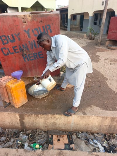 This image captures an outdoor scene in Alizaga, Nigeria, centered around a water collection point. An adult male, dark-skinned, wearing a light-colored long-sleeved tunic and matching trousers with dark flip-flops, and sunglasses pushed onto his head, is bent over. He is pouring water from a small, dirty white bucket into a large, silver-colored metal bowl, creating splashes. Adjacent to him is a large, rusty red metal tank with "BUY YOUR CLEAN WATER HERE" stenciled in black letters on its side, along with the characters "Y4S". A spigot is visible on the tank. Two orange/yellow plastic jerry cans are on the ground near the tank, one fitted with a purple plastic funnel. The immediate ground around the water collection area is unpaved, wet, and muddy. A concrete curb separates this area from a shallow ditch in the foreground, which is heavily filled with assorted refuse, including plastic bottles, bags, and a flattened cardboard box branded "MONSTER ENERGY". In the background, various residential or commercial buildings with cream, blue, and light green paint are visible, along with sparse vegetation and what appears to be a motorcycle.