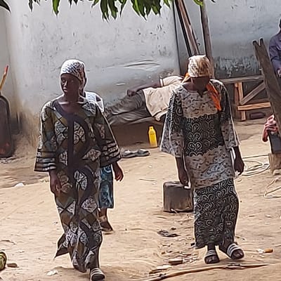 The image captures an outdoor scene featuring several individuals on sandy ground, likely in a village or rural setting. The image is oriented vertically (rotated 90 degrees clockwise).
In the foreground, a woman stands on the right, facing slightly away from the viewer, with her gaze directed downwards. She is wearing a long, patterned dress or robe with motifs in shades of green, blue, white, and brown. Her head is covered with a patterned white headscarf, and she has dark sandals with light-colored soles on her feet.
Behind her, slightly to the left, a man stands wearing a light-colored, possibly cream or beige, short-sleeved shirt and dark pants. His face is not clearly visible. To the man's right, a second woman stands, wearing a long dress or robe with a dark base and intricate white geometric patterns. She has a light-colored headscarf covering her head and an orange piece of fabric draped over her neck or shoulders. She is also wearing dark sandals with white straps.
On the sandy ground, near the man and the second woman, a bright yellow plastic bottle lies horizontally. A dark, possibly metal or plastic, container or pot is also visible near the feet of the second woman. The ground itself is uneven and appears to be made of sand or dirt, with some scattered small debris.
In the background, on the left side, a simple, light-colored building or wall is visible. Further back and to the right, various wooden structures, possibly parts of furniture like a table or support beams, can be seen. In the top left corner, a person is partially visible, appearing to be seated near some wooden structures. Green foliage from a plant extends into the frame from the far left side. The scene is brightly illuminated, suggesting it is daytime. No text is visible in the image.