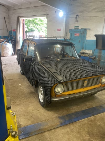 A modified yellow sedan, possibly a Lada, is parked inside a garage in Laishevo, Russia. The car's exterior, including the hood and sides, is covered in a black textured material with numerous small protrusions, resembling a form of automotive armor or protection. The car also has a roof rack. The garage appears to be a workshop, with tools, machinery, and a large bag of material visible in the background. A blue door with Russian signage indicating "EXIT" and another sign about "CARE" is present on the back wall. The floor is concrete, and a yellow car lift is positioned in the foreground. The scene is lit by overhead lighting and daylight streaming through an open doorway.