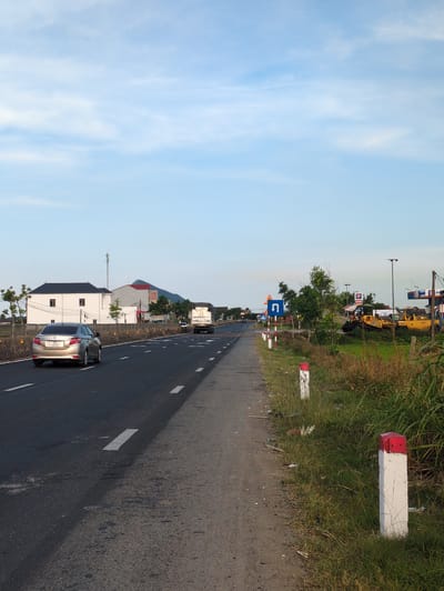 The image depicts a rural road in Kỳ Anh Commune, Vietnam, under a partly cloudy sky. A paved road with white lane markings runs diagonally from the lower left to the upper right of the frame. A silver sedan, bearing the license plate "38A 179.67", is traveling on the road, in the left lane. A white truck is seen further down the road, also in the left lane.
To the left of the road, a low concrete barrier separates the road from a grassy verge and a white building with a dark roof, featuring several windows. Beyond the building, a white tower with antennas is visible. In the background, a hill with a red-capped peak is partially obscured by trees.
To the right of the road, a narrow, unpaved shoulder is lined with grass and vegetation. Several red-and-white striped posts are spaced along the edge of the shoulder, marking the road's boundary. Further to the right, a gas station with a blue and red sign is visible, alongside two yellow bulldozers parked on a grassy area. The overall scene suggests a transition zone between rural countryside and infrastructure development.