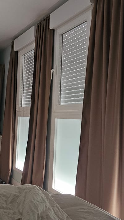 The image shows two windows with white frames and brown curtains partially drawn. The windows are fitted with roller shutters, which are partially open, allowing light to filter through. The lower part of the windows appears to be frosted glass. In the foreground, a white duvet is visible, suggesting the photo was taken from a bed. The scene appears to be indoors, possibly a bedroom, with the windows providing natural light. The location is identified as María De Huerva, Spain.