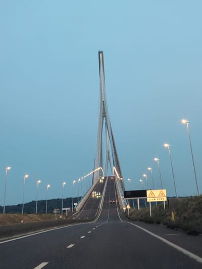 The image shows a view from a vehicle traveling on a multi-lane highway that leads onto a large cable-stayed bridge. The bridge has a prominent central pylon with cables supporting the roadway. The sky is a clear, pale blue, suggesting dusk or dawn. Streetlights are illuminated along the sides of the highway and bridge, casting a warm glow. Several vehicles are visible on the road, some ahead and some approaching. On the right side of the road, there are traffic signs. One sign displays two yellow triangles with figures of people walking, and the text "SLOW DOWN" below. The surrounding landscape on the left appears to be a grassy embankment with trees in the distance. The location is identified as the Pont de Normandie in Sandouville, France.