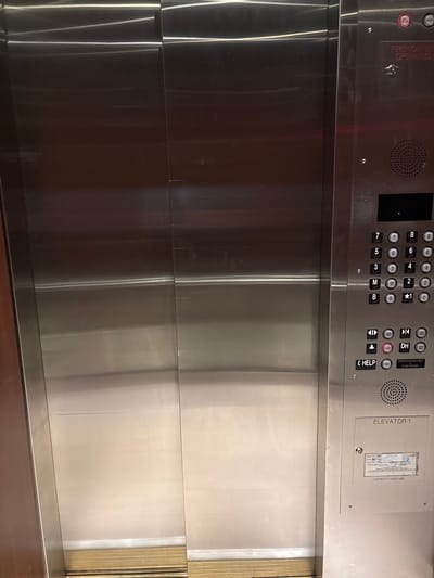 The image shows the interior of an elevator, specifically the control panel and the elevator doors. The control panel is made of brushed metal and features a keypad with buttons numbered 1 through 8, as well as buttons for M (Mezzanine), B (Basement), and ★1 (likely a special floor). There are also buttons for door open, door close, up arrow, down arrow, alarm, and help. Above the keypad, a digital display shows a green light, indicating the elevator's status. A small speaker is located below the keypad. To the right of the control panel is a framed inspection certificate. The elevator doors appear to be made of polished stainless steel, reflecting the surroundings. The floor of the elevator is visible at the bottom, with a metallic threshold. There is no indication of the city of San Francisco in the image, and no people or actions are depicted. The environment is clean and sterile, typical of an elevator interior.