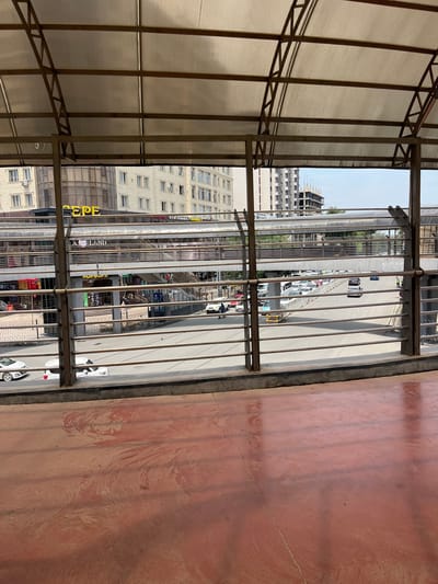 The image shows a view from an elevated walkway or bridge in Osh City, Kyrgyzstan. The foreground is a reddish-brown concrete surface with some dirt and debris. Behind this, metal railings form a barrier, beyond which a street level is visible. Several cars, including white sedans and a black SUV, are driving on the street. Further back, there is an elevated pedestrian walkway or bridge with a translucent roof, where people can be seen walking. Buildings, some appearing to be multi-story residential or commercial structures, line the street and the walkway. One building in the middle ground has a prominent yellow sign with the Cyrillic letters "EPE". Another building to the left displays the word "LAND" in English. Underneath the pedestrian walkway, there are shops or commercial establishments with various signs, some in Cyrillic script. In the background, a partially constructed building with visible scaffolding suggests ongoing development. The sky is mostly clear with some clouds. The overall scene depicts an urban environment with active traffic and pedestrian movement.