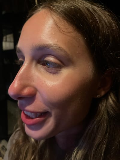 The image is a close-up, side profile of a young woman's face, taken in Kertasari, Indonesia. Her brown hair is partially visible, framing her face, which is illuminated by warm light that highlights the sheen of her skin. She is wearing light-colored lipstick and her mouth is slightly open, revealing her teeth in a subtle smile. Her right eye is visible, with a green iris. She has a small diamond stud earring in her right ear. The background is dark and indistinct, suggesting an indoor setting with low lighting conditions.