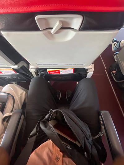 The image shows a first-person perspective of a person sitting in an airplane seat, with their legs extended forward and a black bag resting on their lap. The seat is a typical airplane seat with a tray table in front, which is partially visible. The tray table has labels indicating "Fasten Seat Belt While Seated" and "Life Jacket Under Your Seat". There are also two red and yellow safety information cards tucked into a pocket on the tray table. The seats are upholstered in red and black, and the floor is a dark red carpet. The person is wearing black pants and black sneakers with white laces. In the background, another passenger's legs and feet, wearing sneakers, are visible. The overall environment suggests an airplane cabin. The image was taken in Mai Khao, Thailand.