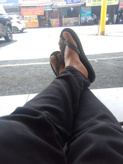 The image shows a person's legs and feet, wearing black pants and flip-flops, resting on a white ledge with a road in the background. Behind the person, there are several shops and stalls, indicating an urban or commercial setting. One shop has a sign that reads "BERKAH SUNDA" with offerings like "AYAM KREMES, MIE GORENG, NASI GORENG, KWETIAU". Another sign advertises "TIK TOK LAUNDRY" with a price of "RP 7.000 KILO". Various vehicles, including cars and motorcycles, are parked or moving in the background. The overall environment appears to be a busy street in Cianjur, Indonesia, likely a commercial area with food vendors and service establishments. The lighting suggests it is daytime.