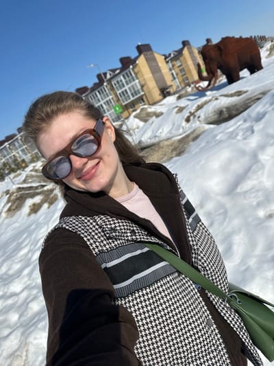 A young woman is captured in a close-up selfie, positioned in the foreground and smiling directly at the camera. She wears brown-framed reflective sunglasses, a dark brown hooded jacket, a houndstooth-patterned vest with black and white horizontal stripes across the chest, and a light pink undershirt. A dark green shoulder bag is visible across her body. The ground is covered in significant amounts of white and partially melted, dirty snow. In the middle ground, to the right of the woman, a large, brown sculpture of a woolly mammoth with tusks stands on a snow mound. The background features several multi-story residential buildings with beige facades, multiple windows, and dark roofs, under a clear blue sky. A street light and a partially visible green sign are also present near the buildings. The scene is located in Новое Шигалеево, Russia.