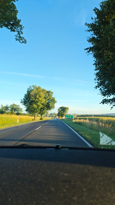 The image is a photograph taken from the interior of a vehicle, looking out over a highway. The road stretches ahead, flanked by green fields and trees. The sky is a clear, bright blue. The time of day appears to be late afternoon, given the long shadows cast by the trees and the warm sunlight. In the distance, a green road sign indicates the direction to Legnica, suggesting the location is near Stanowice, Poland. There are no people visible in the image, and the focus is on the natural scenery and the road itself. The overall impression is one of a peaceful, rural journey.