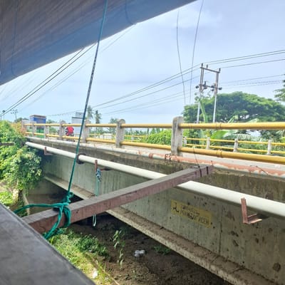 The image shows a bridge in Mesjid Tuha, Indonesia. The bridge has concrete pillars supporting a roadway, with yellow railings on either side. There are power lines overhead, and lush green vegetation on the left side of the bridge. A person on a red motorcycle is visible crossing the bridge. The underside of the bridge is also visible, showing concrete beams and pipes. There is a sign on the underside of the bridge with the text "23MP0020 PRaktAK ACEH". The ground beneath the bridge is a mix of soil and vegetation.