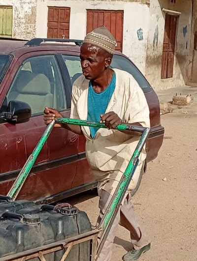An adult male is depicted in a daytime street scene in Gayawa, Nigeria. He possesses dark skin, a shaved head covered by a light-patterned skullcap, and a visible lesion or scar on his forehead. He is wearing a light-colored tunic over a teal t-shirt, light trousers, and dark footwear. His expression is strained, mouth slightly open, as he grasps the green-painted metal handles of a two-wheeled pushcart. The cart carries at least two black plastic containers, likely jerrycans, one of which has a reddish-brown cord wrapped around its cap. Behind the man and cart, a maroon sedan with a roof rack is parked. In the background, two weathered, light-colored buildings with brown window frames and some faded markings are visible. The ground is unpaved, appearing as dry earth.