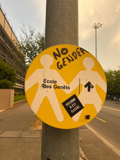 A circular yellow sign with white silhouettes of two people is attached to a gray cylindrical pole. The silhouettes are connected by the hands, and one has a white arrow pointing upwards in the center of its torso. The text "NO GENDER" is written in black, handwritten letters above the silhouettes. Below the silhouettes, the text "Ecole des Genêts" is written in black text. A small black square sticker with white text reads "PERSONNE N'EST ILLEGAL" and is placed on the lower right side of the sign. At the bottom right of the sign, a city crest for "VILLE DE GENÈVE" is visible. The background shows a building with multiple floors on the left, trees, a street with yellow lines, and a lamppost in the distance. The sky is overcast with a warm hue.