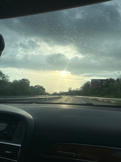 The image shows a view from inside a car, likely moving along a highway near Kipra, Bulgaria. Raindrops are visible on the windshield, partially obscuring the outside scene. The sky is overcast with dark clouds, but the sun is breaking through, creating a bright glare. On the right side, the car's dashboard is visible, indicating it's an Audi with a "quattro" emblem. Outside, trees line the highway. A sign can be seen in the distance.