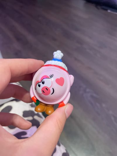 A person's hands are holding a small, pink, pig-shaped toy. The toy has a blue and white hat with a pom-pom, a red trim around its head, and red accents on its feet and arms. It has large cartoon eyes, a pink nose, and two pink heart shapes on its cheeks. The toy is resting on a surface with a black and white pattern, possibly a rug or fabric, in front of a wooden floor. The background is blurred and out of focus. There is no identifiable location in the city of Vaganovo Massiv, Russia.