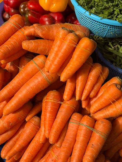 This image captures a market stall in Jakande, Nigeria, displaying a variety of fresh produce. The foreground is dominated by a dense pile of numerous orange carrots, many of which are grouped into bundles of three to five, secured with yellow or green rubber bands. In the upper left background, an assortment of red peppers, including bell peppers and other darker red varieties, is visible, along with one yellow bell pepper. To the upper right, a light blue perforated plastic basket holds a quantity of green leafy herbs. A darker blue container, partially visible below the herbs, contains green beans or similar elongated green vegetables. The scene is a static arrangement of produce, with no individuals or active interactions present.