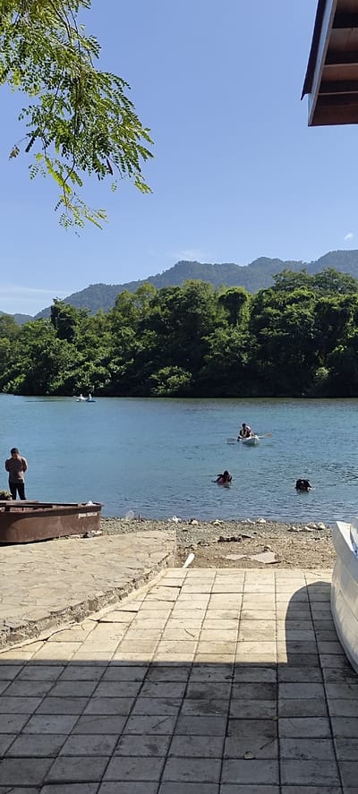 In Lamseunia, Indonesia, a scenic view unfolds on a clear day. A calm, blue river dominates the scene, with lush green mountains forming a backdrop. The foreground features a paved area leading down to a rocky shore, with a rustic brown boat beached on the left. A man in a dark shirt stands by the boat, looking towards the water.

Several people are enjoying the river. Two individuals are kayaking in the distance, while closer to the shore, three people are swimming, two with their heads visible above the water. The water is clean and inviting, reflecting the bright blue sky. The overall atmosphere is one of tranquility and outdoor recreation.