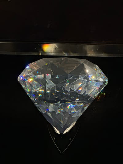 The image shows a large, faceted, clear crystal object resembling a diamond. The crystal is positioned centrally against a black background. Light is refracting through the crystal, creating a spectrum of rainbow colors on its facets and in the surrounding area. The crystal appears to be displayed in a dark environment, possibly a display case or a darkened room, given the reflections seen at the top of the image. The specific location in Wattens, Austria, is not discernible from the image itself, but the presence of such a crystal would likely indicate a museum, gallery, or shop related to crystal production or display, given Wattens is known for Swarovski Crystal Worlds. The scene is static and focuses solely on the object and its interaction with light.