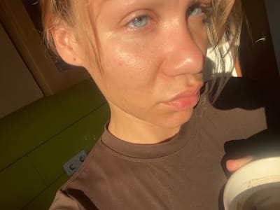A close-up, waist-up portrait of a young blonde woman with blue eyes is captured in the image. She is positioned in the foreground, looking directly at the camera with a neutral expression. The woman is wearing a dark brown t-shirt. To her left, a wall with green tiles is visible, featuring a white double electrical outlet. The lighting suggests an indoor environment, possibly a room with sunlight streaming in, creating strong shadows on the right side of the frame. The woman is holding a white cup in her right hand, which is partially obscured. The overall scene is informal and personal.