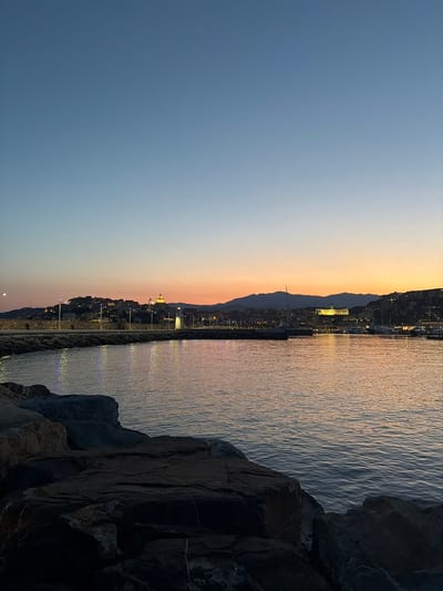 The image depicts the harbor of Imperia, Italy, during the twilight hours. In the foreground, dark, rugged rocks are visible, leading to the calm waters of the harbor. The water reflects the soft colors of the sunset, with hues of orange, pink, and blue blending in the sky.
The middle ground features a stone pier lined with streetlights that cast a warm glow on the water. Beyond the pier, the city of Imperia is illuminated by numerous lights, creating a picturesque silhouette against the darkening sky. A prominent church with a golden dome stands out on the hillside, a recognizable landmark of the city.
In the background, the gentle slope of the hills surrounds the harbor. The sky transitions from a clear, pale blue at the top to a warm gradient of colors near the horizon, indicating the end of the day. A few boats are moored in the harbor, their masts subtly visible against the illuminated cityscape. There are no people clearly visible in the frame. The overall atmosphere is serene and tranquil, capturing the beauty of a coastal Italian city at dusk.