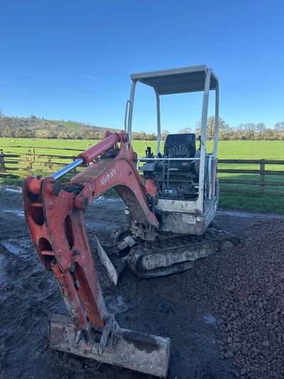 A red and white Kubota mini excavator is stationary on a rural property in Broad Chalke, United Kingdom, under a clear blue sky. The machine's hydraulic arm, labeled "Kubota," is extended downwards, with its dirt-encrusted bucket resting on dark, wet mud in the foreground. The excavator's track system and lower chassis are heavily soiled with mud. A black operator's seat is visible beneath a white protective canopy. To the right of the machine, the ground transitions to a dark, coarse gravel path. In the midground, a wooden fence separates the immediate area from a vibrant green pasture where several white sheep are grazing. The background features rolling green hills partially covered with deciduous trees. No individuals are present in the scene.