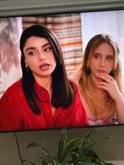 The image shows a close-up of two young women on a television screen. The woman on the left has dark hair and is wearing a red shirt. She is looking to her left with her mouth slightly open. The woman on the right has blonde hair and is wearing a floral top. She is resting her chin on her hand and looking to her left. The background is blurred, but appears to be a room with striped curtains or a tapestry. The bottom of the image shows the top of a television set and a plant with green leaves. The image is taken in the city of Springboro, United States of America.