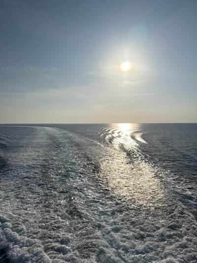 The image depicts an open sea environment near Kamariotissa, Greece, captured from a vessel moving forward. The scene is dominated by a clear sky in the upper half and the sea in the lower half. The sun is a prominent, bright yellow-white disc in the upper right quadrant of the sky, casting a strong glare and producing a reflective path of shimmering light across the water's surface, extending towards the lower right. The sky is light blue, transitioning to a lighter shade near the horizon, with a faint linear contrail visible horizontally in the mid-upper section. A small, dark, distant object, possibly a bird or aircraft, is visible in the upper left sky. The sea exhibits a dark, deep blue-grey hue. A wide, turbulent wake of white foam and disturbed water trails from the immediate foreground towards the horizon, indicating the recent passage of a vessel. No people or other distinct subjects are visible beyond the natural elements and the effects of a moving vessel.