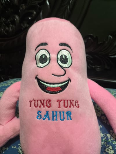 The image shows a close-up of a pink plush toy with a smiling face. The toy has embroidered features, including large eyes with black outlines and white and green centers, eyebrows, a nose, and a wide mouth with red lips and black teeth. The words "TUNG TUNG SAHUR" are embroidered on the front of the toy in red and blue lettering. The toy is positioned against a dark, ornate background, possibly furniture, and rests on a blue and green patterned fabric. The context provided by "Ujongreuba, Indonesia" suggests the image was taken in that location.