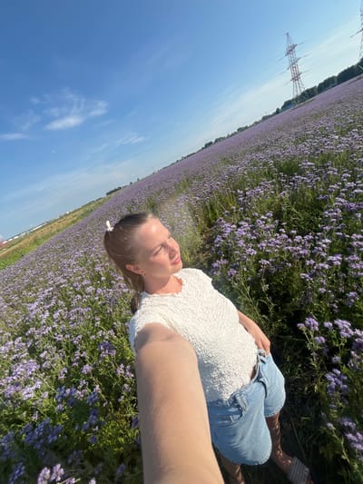In Sovkhoza "Pyatiletka", Russia, a young woman is captured in a selfie, standing in a vast field of blooming purple flowers under a clear blue sky. She is wearing a textured white top and light blue denim shorts, with her hair tied back in a ponytail. Her right arm is extended to take the photo, and she is looking off to the side, bathed in sunlight. In the background, power lines and transmission towers are visible, contrasting with the natural beauty of the floral landscape. The image is taken from a low angle, emphasizing the expanse of the field and the towering sky.