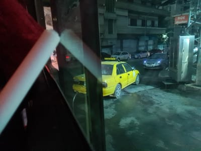 An upside-down urban scene is visible. A yellow car dominates the view, accompanied by other vehicles on a street lined with buildings. A sign labeled "Alsultan" is partially visible. The image is framed by a dark, abstract foreground with what appears to be a structural element, possibly a pole or support beam. The perspective is unusual, giving the impression of looking at a reflection or an inverted reality.