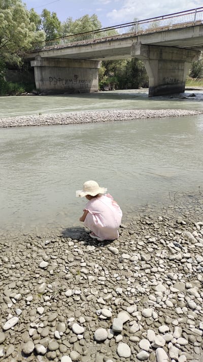 A young child wearing a straw hat and a pink dress is crouching on a rocky shore, playing at the edge of a river. The child is facing away from the camera, toward the water. In the background, a concrete bridge spans the river. Graffiti is visible on the bridge's support pillars. Trees and vegetation line the riverbanks and the area beyond the bridge. The sky is cloudy. This scene is located in Taldykorgan, Kazakhstan.