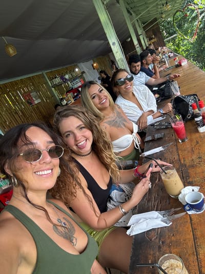 A group poses at a long wooden table. The image, taken from the end of the table, features four women in the foreground smiling and looking directly at the camera. Some sport sunglasses. Behind them, several other people sit, also looking toward the camera. Drinks, tableware, and personal items are scattered on the table. A bamboo structure and tropical foliage form the background, suggesting an outdoor setting.