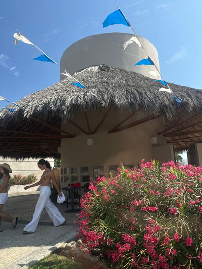 The image shows a brightly lit outdoor scene in Acapulco de Juárez, Mexico. A circular structure with a thatched roof dominates the left side of the frame. White and blue triangular pennants are strung along the perimeter of the roof. Adjacent to the structure, on the right, is a building with a tan exterior and multiple small, square windows. A large flowering bush with magenta blossoms is beside the building.
Two women are visible on a paved area in the foreground. One woman wears a bikini top and white pants, holding a white garment. Another woman is near the bottom of the frame, lying on the ground and wearing a sun hat. Outdoor furniture is visible in the background, including a table and chairs.