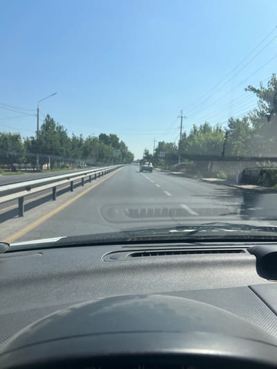 A first-person perspective shot from inside a vehicle captures a straight, paved road with a clear blue sky overhead. Trees line the right side of the road, and a guardrail is visible on the left. The road is marked with a yellow line on the left and white dashed lines in the center. A pedestrian crossing sign is posted on the right side of the road. Two vehicles are visible on the road ahead, one in the distance and another closer. The dashboard of the vehicle in the foreground is visible, with a textured gray surface and a speaker grille. The image was taken in Uymavut, Uzbekistan.