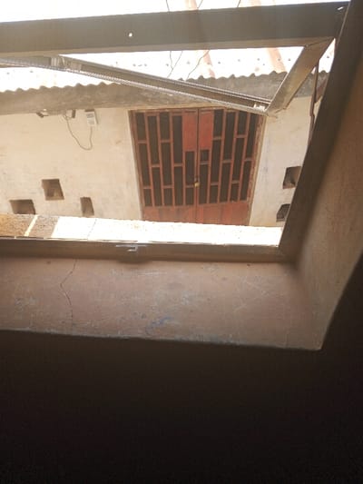 The image presents an exterior scene in Oko Erin, Nigeria, viewed from an elevated, partially shaded perspective. In the immediate foreground, a tan-colored concrete ledge occupies the lower third of the frame, displaying surface cracks and minor abrasions. Beyond this ledge, a brightly illuminated courtyard or open area is visible, bordered by light-colored, possibly rendered, walls.
Centrally located within the opposing structure is a reddish-brown double door, constructed with a grid-like pattern of rectangular panels. A silver-colored padlock secures the right door leaf. To the left of the door, a white rectangular electrical device or keypad is mounted on the wall, with a black electrical cable descending from it. Both the wall sections flanking the door feature multiple small, square-shaped cutouts. Above the walls, a corrugated metal roof is visible, partially obscured by dark-colored structural metal beams that span the upper portion of the frame. The scene is static and devoid of human presence or active interaction.