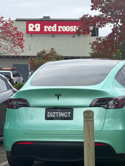 A light green Tesla Model Y, with the license plate "DIZTINCT," is parked in front of a Red Rooster restaurant in Bassendean, Western Australia. The car is positioned near a wooden post or barrier. Red-leaved trees are visible in the foreground and surrounding the building. Other vehicles are partially visible in the background, further down the street. The sky is overcast.