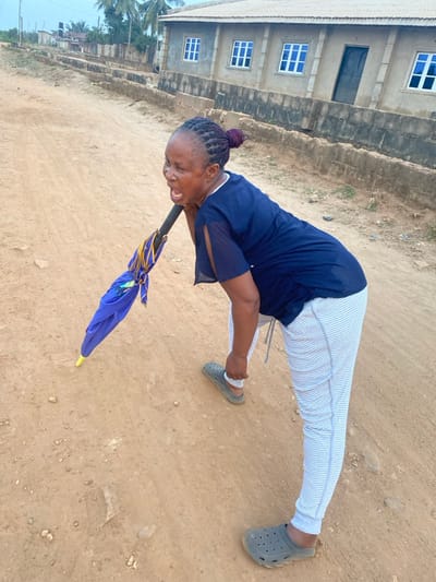 An adult female is depicted bent forward at the waist on an unpaved, reddish-brown dirt road in Odo Ona, Nigeria. Her mouth is open, appearing to scream or shout, and her eyes are wide. She has dark braided hair pulled into a reddish-purple bun. She wears a dark blue short-sleeved top with split sleeves, light-colored horizontally striped pants, and grey Crocs-style shoes. Her right hand grasps the handle of a folded blue umbrella with a yellow tip, which rests on the ground beside her. Her left hand is positioned near her left hip. In the background, a single-story building with a light-colored stucco-like facade, a corrugated metal roof, multiple rectangular windows with blue frames and white grilles, and a dark entrance door is visible. A low, stepped concrete or stone wall runs along the side of the road in front of the building. Further in the background, more structures and green foliage are discernible under an overcast sky.