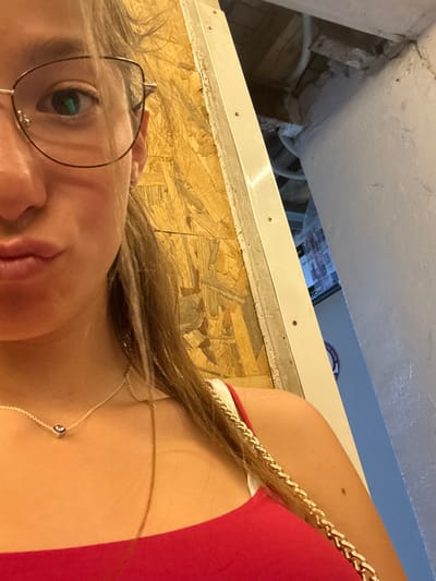 The image shows a close-up of a young woman with blonde hair, wearing glasses and a red top. She is wearing a delicate silver necklace with a heart-shaped pendant. The background is a textured wall with wood paneling, and a glimpse of a doorway or opening reveals an interior space with exposed pipes and a blue wall. Based on the provided information, the location is Bozhurishte, Bulgaria. The image appears to be a selfie taken by the woman.