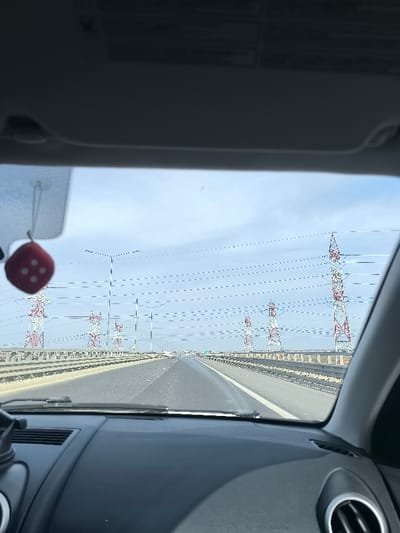 The image depicts a daytime view from the interior of a vehicle traveling on a multi-lane highway, specifically located within or near Glina, Romania. The perspective is forward through the windshield, showing the vehicle's dark gray dashboard in the lower foreground. A red fuzzy dice, marked with white spots representing the numbers "4" and "3," hangs from the rearview mirror assembly on the upper left, slightly out of focus. Part of a silver-rimmed circular air vent is visible in the lower right corner of the interior.
Outside the vehicle, the highway extends straight into the distance under a light blue sky with scattered thin clouds. The road features multiple lanes and appears to be a divided highway, possibly an elevated section or a bridge, flanked by concrete barriers and metal guardrails. Prominent in the midground and background are several tall, red and white lattice transmission towers (power pylons) positioned on both sides of the highway. Numerous power lines are strung horizontally across the sky, connecting these towers. Slender street light poles are also visible along the roadway. No individuals are discernible outside the vehicle.