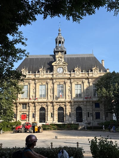 The image depicts the Hôtel de Ville (City Hall) of Ivry-sur-Seine, France, on a clear, sunny day. The ornate architecture of the building, with its detailed carvings and a prominent clock tower, is visible. Trees frame the scene, partially obscuring the building.

In front of the Hôtel de Ville, several tractors are parked on a paved area. One is red, another is yellow, and a third, partially obscured, is also visible. A person wearing a helmet is seated on a bench in the foreground, looking towards the building. Other individuals are seen in the background, some interacting near a park or plaza area. The presence of the tractors suggests a potential event or demonstration.

Visible text includes "FRATERNITE" and "LIBERTE" above doorways on the building, which are French words for "Brotherhood" and "Liberty" respectively, aligning with the national motto of France. The sky is a vibrant blue, indicating good weather and likely daytime. The sunlight casts shadows, suggesting it is mid-day or early afternoon.