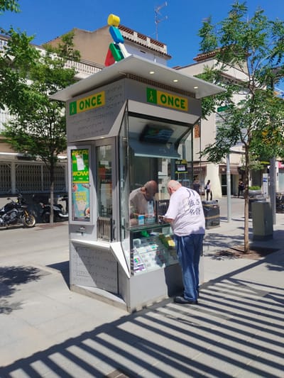 The image shows a vendor stand in Rubí, Spain, with a man standing in front of it examining something inside. The stand is marked with "ONCE," and its exterior is adorned with braille. Colorful, stacked geometric shapes are on its roof. The stand offers various items on display, with promotional posters visible on its sides.
The man is wearing a white t-shirt with Japanese characters and blue jeans. Inside the stand, a reflection of a bald-headed man can be seen. The surroundings indicate an urban environment with buildings, trees, motorcycles parked nearby, and street furniture like waste bins. The scene is bathed in sunlight, casting distinct shadows on the ground. A street sign and multiple storefronts are visible across the street behind the vending stand.