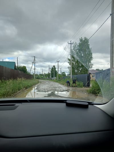 The image depicts a scene in Verkhneye Dubrovo, Russia, on a cloudy day. The perspective is from inside a vehicle, looking out through the windshield. The road is unpaved and heavily waterlogged, with numerous large puddles and muddy patches. Two individuals are visible on the right side of the road, appearing to be engaged in manual labor, possibly digging or clearing debris with shovels. They are dressed in casual clothing. In the background, there are residential buildings, fences, and numerous utility poles with overhead wires, characteristic of a rural or semi-rural area. Lush green vegetation lines the sides of the road. The sky is overcast with heavy, grey clouds.
