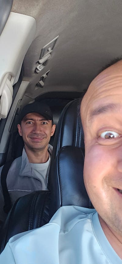 The image shows two men in a car. The man in the foreground is a bald man with his face very close to the camera, his right eye is visible and his mouth is open in a smile. The second man is in the background, wearing a black baseball cap and a grey jacket over a white shirt. He is smiling and looking directly at the camera. The interior of the car is visible, including the ceiling with lights and the seats. The overall scene suggests a casual moment captured during a car ride. The question of this image being taken in Guzar, Uzbekistan is not answered by the image itself, as there are no identifying landmarks or contextual clues.